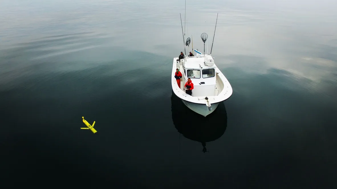 Aerial view of the recovery boat approaching a surfaced glider on glassy water.