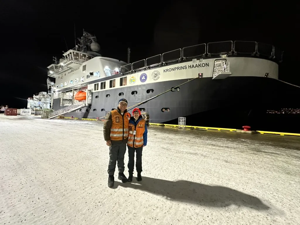 Two lab members in front of the Norwegian icebreaker R/V Kronprins Haakon at a snowy dock in northern Norway.