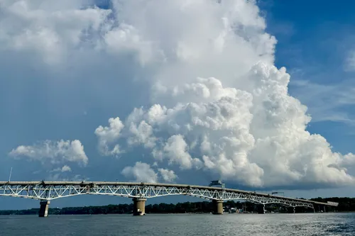 Towering cumulus building over the York River on a summer afternoon, seen from the water, with the Coleman Bridge crossing the frame beneath it and Yorktown on the far shore.